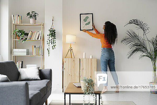 Junge Frau hängt Bilderrahmen an Wand im Wohnzimmer zu Hause