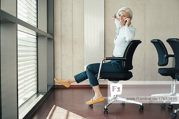 Glückliche Geschäftsfrau  die auf einem Stuhl im Büro sitzt und mit ihrem Smartphone spricht