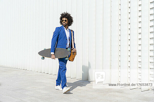 Businessman holding skateboard and shoulder bag walking on footpath