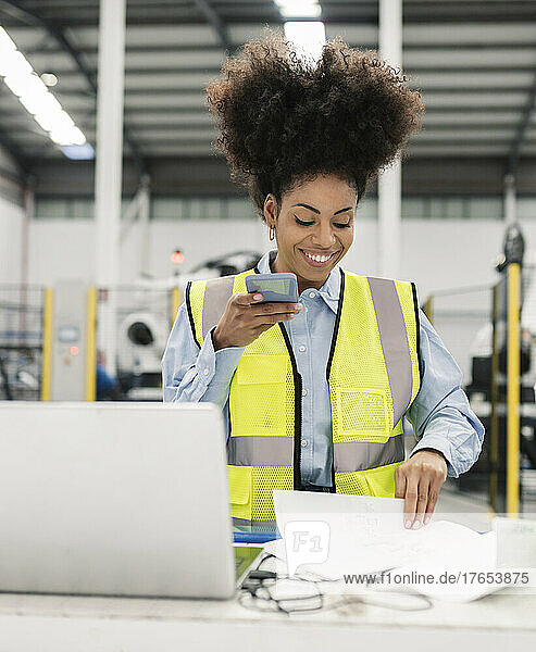 Smiling engineer sending voicemail through smart phone analyzing documents in factory