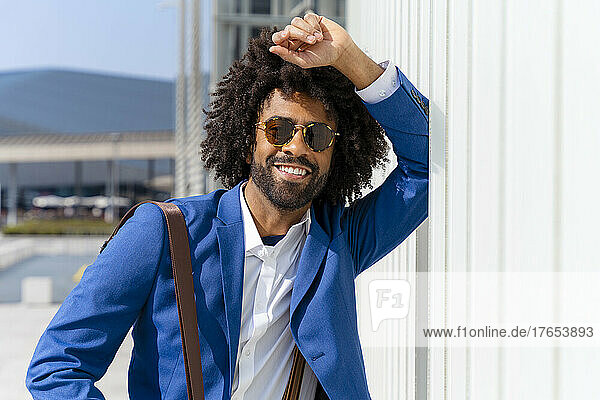 Smiling businessman with curly hair standing by wall on sunny day
