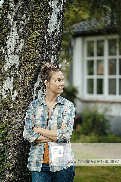 Woman standing with arms crossed by tree trunk at backyard
