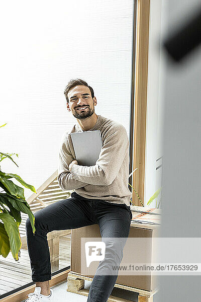 Smiling man sitting on box holding tablet PC at home