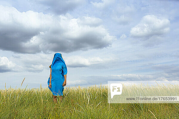 Mit blauem Schal bedeckte Frau steht inmitten von Gras auf einer Wiese