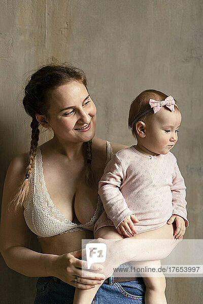 Smiling young mother with cute daughter standing in front of wall