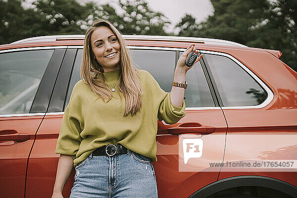 Smiling young woman with key standing in front of car