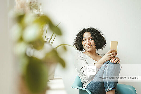 Smiling woman sitting in chair with smart phone at home
