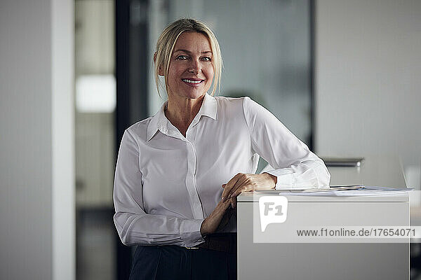 Smiling businesswoman with tablet PC standing by cabinet in office