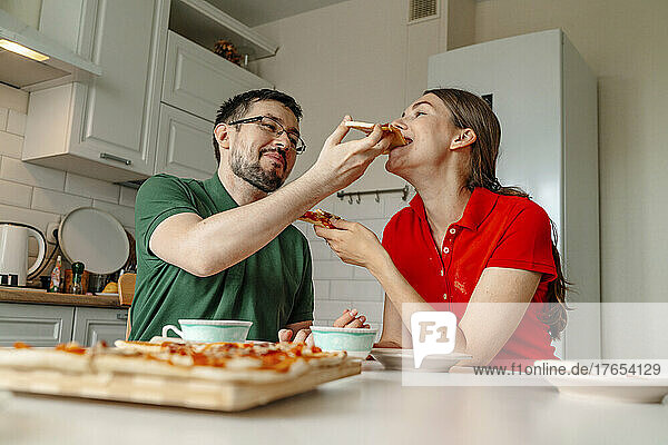 Man feeding pizza to woman sitting at table in kitchen