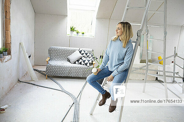 Thoughtful woman with coffee cup sitting on ladder in attic