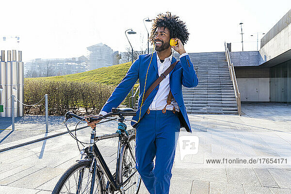 Happy businessman with bicycle listening music through headphones on footpath