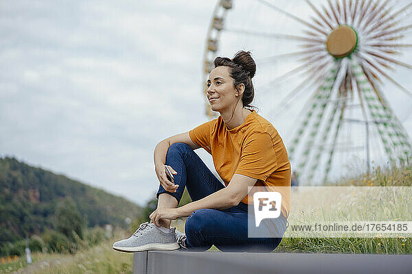 Smiling woman holding smart phone sitting in front of ferris wheel