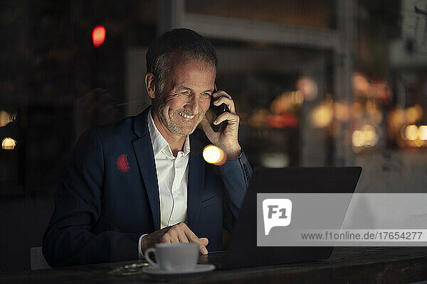 Happy businessman talking on mobile phone and using laptop at night