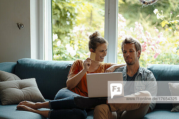 Cheerful woman holding credit card sitting by boyfriend using laptop on sofa in living room