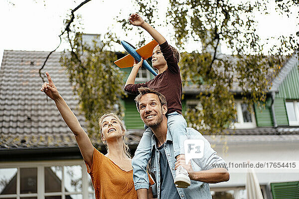 Woman pointing by man carrying playful son on shoulders holding toy airplane in backyard