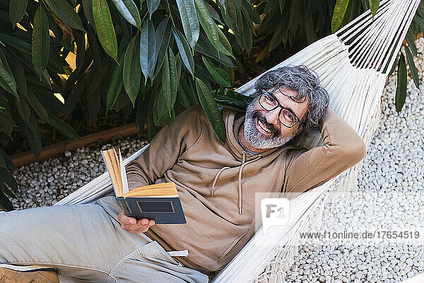 Senior man reading book by lying hammock in backyard