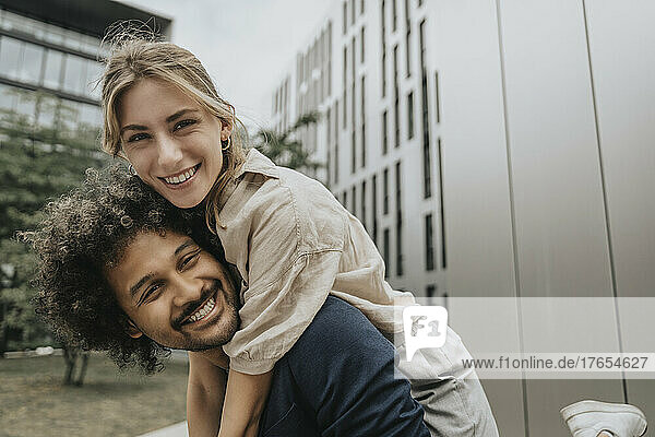 Smiling young man piggybacking girlfriend in front of modern building