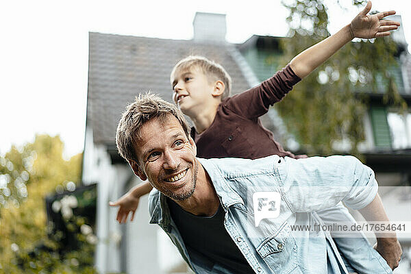 Glücklicher Vater gibt seinem Sohn Huckepackfahrt mit ausgestreckten Armen und genießt es im Hinterhof