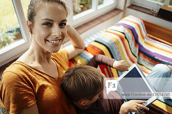 Smiling mother sitting by son using tablet PC on sofa at home