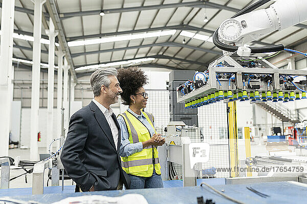 Happy businessman with hands in pockets standing by engineer looking at robotic machine in factory