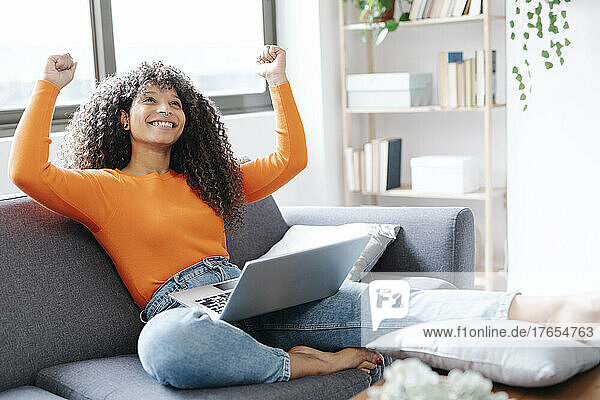 Happy woman gesturing fist sitting with laptop on sofa at home