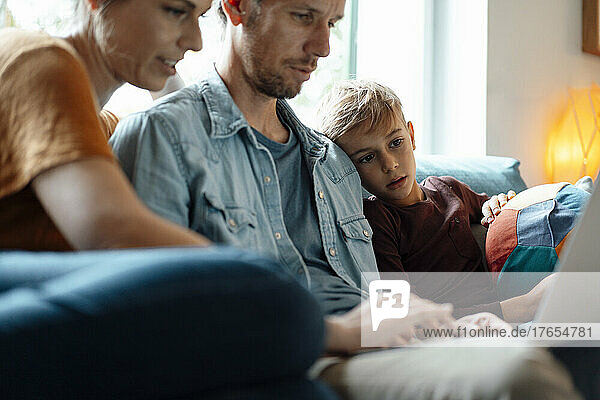 Eltern nutzen Laptop und sitzen zu Hause neben ihrem Sohn im Wohnzimmer