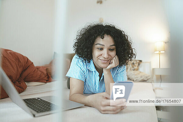 Smiling woman with laptop using smart phone on bed at home
