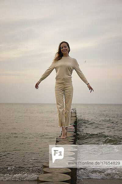 Happy woman with arms raised walking on wooden post at beach