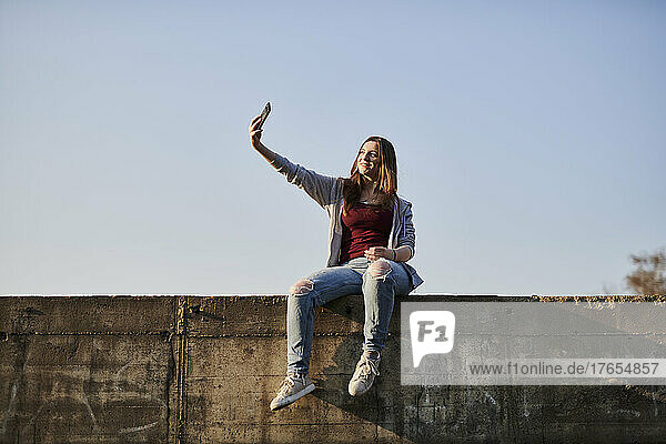 Young woman sitting on concrete wall taking a selfie