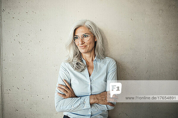 Smiling businesswoman wearing eyeglasses standing with arms crossed in front of wall