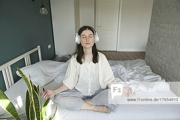 Woman wearing wireless headphones meditating on bed at home