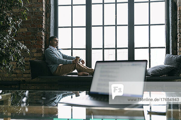 Young freelancer with disposable coffee cup sitting on window sill at home