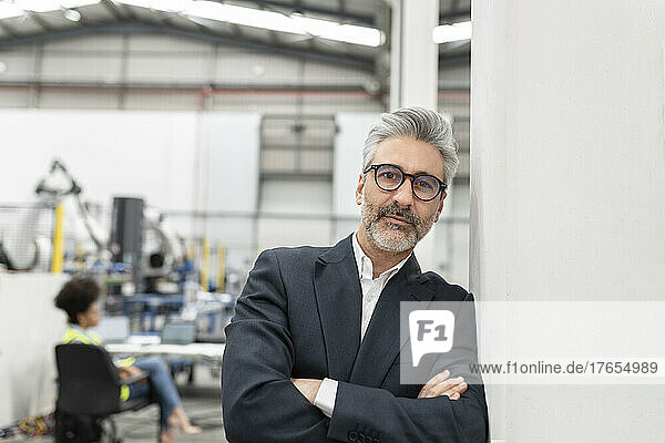 Businessman standing with arms crossed leaning on wall in factory