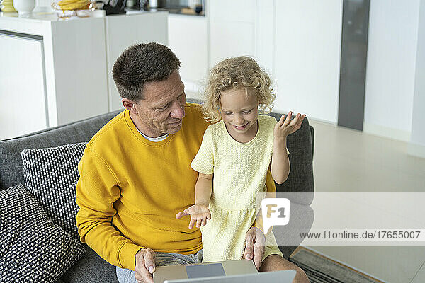 Smiling father and daughter with laptop sitting on sofa at home