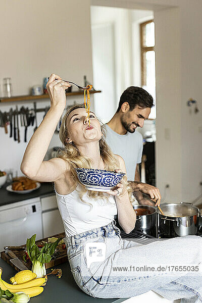 Woman slurping noodles with boyfriend preparing food in kitchen at home