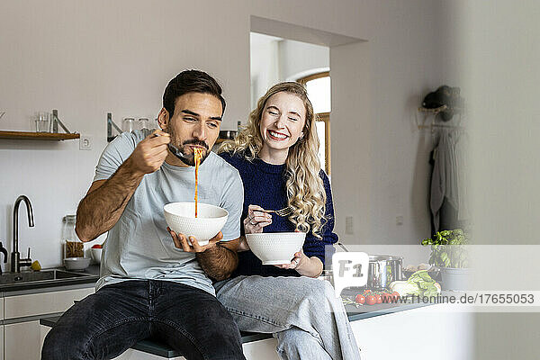 Lächelnde Frau sitzt neben einem Mann und isst Nudeln aus einer Schüssel in der heimischen Küche
