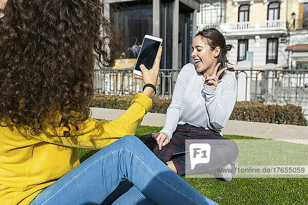 Young woman photographing friend gesturing peace sign through smart phone sitting at park