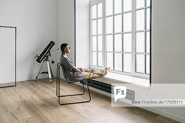 Young man looking through window sitting on chair at home