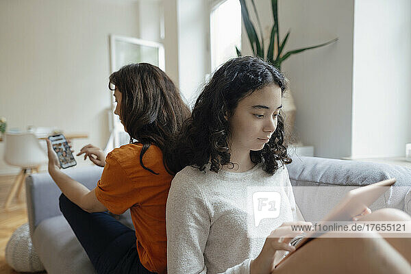 Teenage girl using tablet PC sitting back to back with mother holding mobile phone