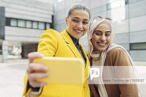 Happy businesswoman taking selfie with young colleague through smart phone