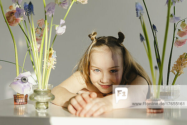Lächelndes Mädchen lehnt auf einem Tisch inmitten von Blumen vor einer weißen Wand