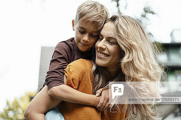 Happy blond woman giving piggyback ride to son at backyard