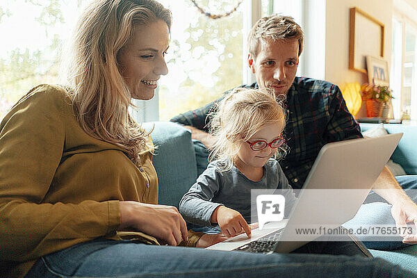 Kleines Mädchen benutzt Laptop und sitzt mit den Eltern zu Hause auf dem Sofa