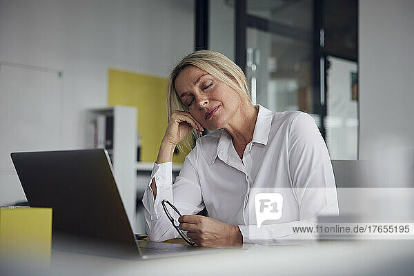 Businesswoman with eyes closed holding eyeglasses with laptop at desk in office
