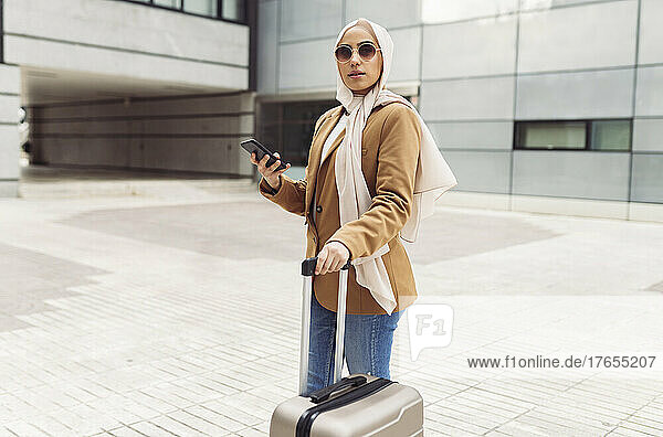 Businesswoman wearing sunglasses holding smart phone standing with wheeled luggage on footpath
