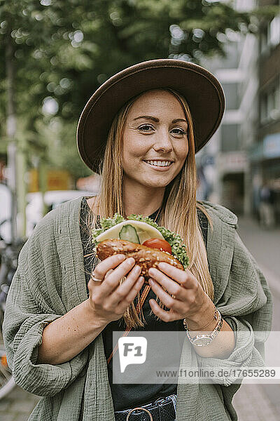 Smiling young woman with sandwich standing on street