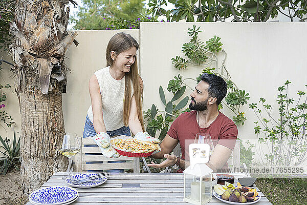 Happy woman showing homemade savory pie to man sitting at table in garden