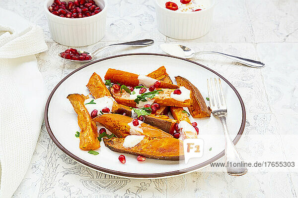 Plate of sweet potatoes with parsley  pomegranate seeds and cumin yogurt lying on white tiles