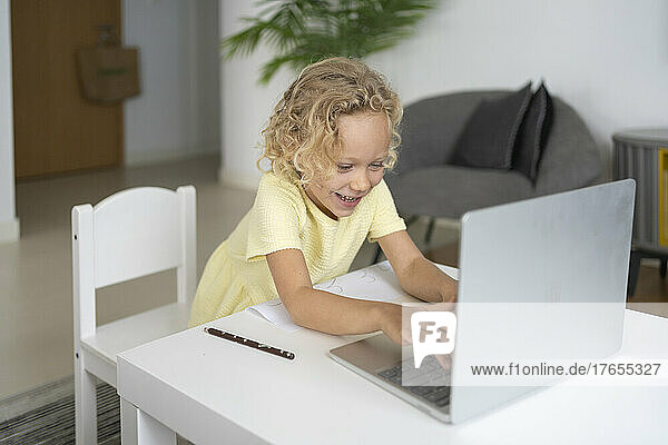 Happy blond girl using laptop sitting at table in living room