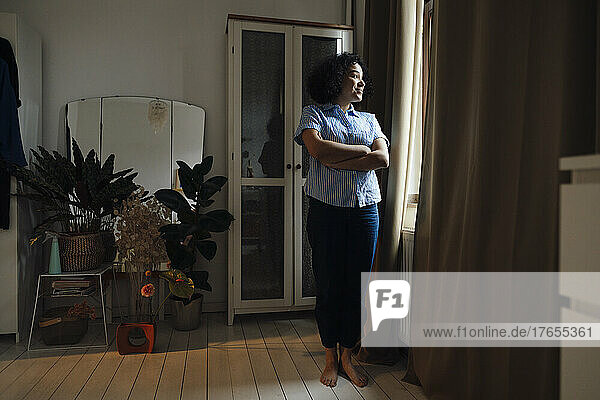 Smiling woman standing with arms crossed looking out through window at home